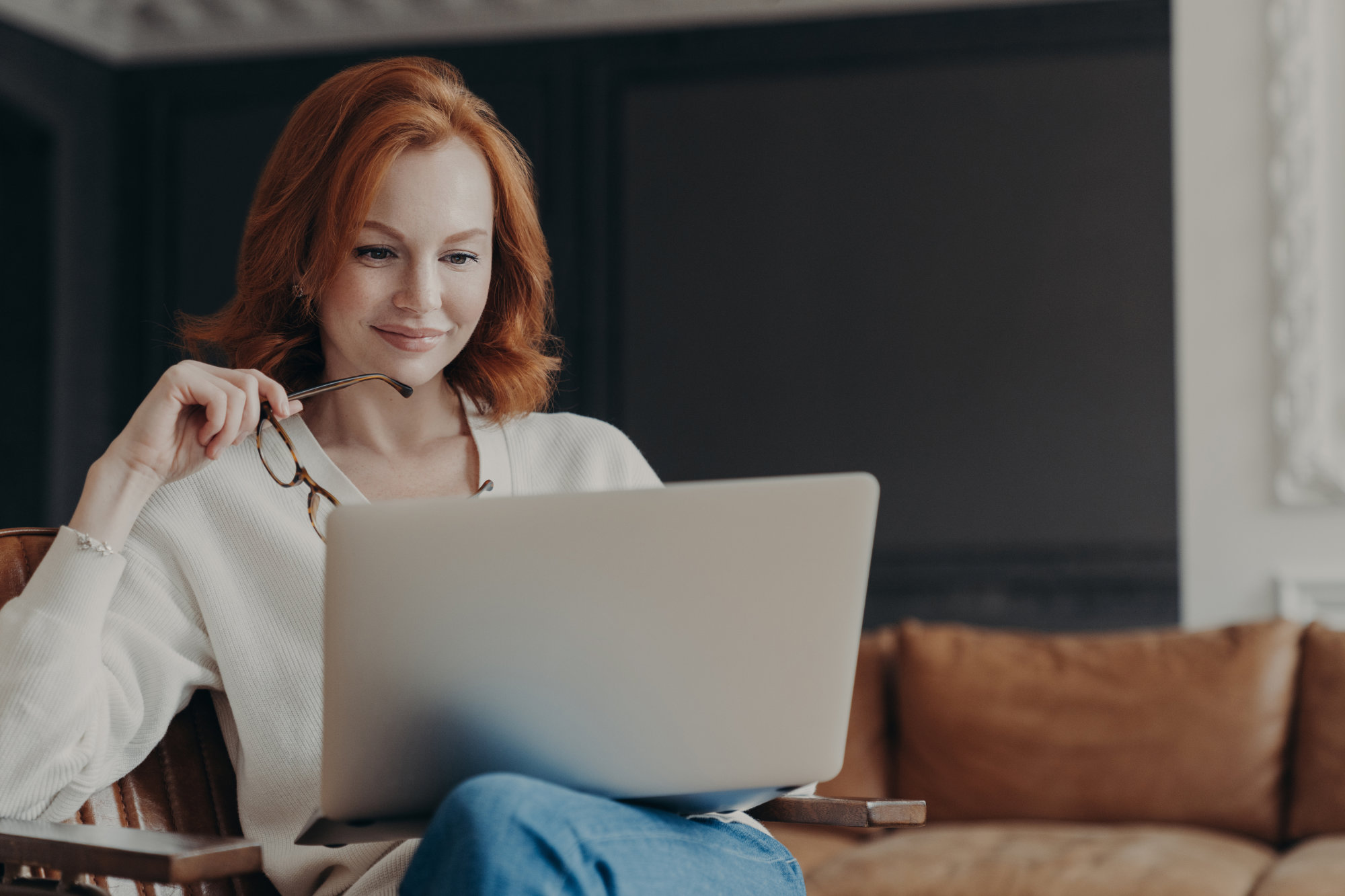 Horizontal shot of happy skilled ginger woman teacher works distantly, checks students works via laptop, holds spectacles, poses in modern cozy apartment. Student checks database for university